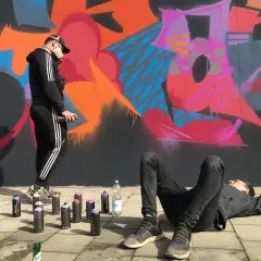 Zwei Künstler, eine Grafikdesignerin und ihr Kollege, sprühen Graffiti. Einer liegt entspannt, der andere sprüht Farbe auf die Wand.