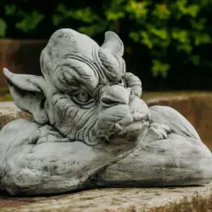Graue Gartenskulptur eines Fabelwesens, das auf einem Holzstamm ruht, mit grünem Laub im Hintergrund.
