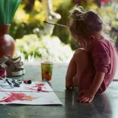 Kleines Mädchen malt mit Pinsel und Farben, um orange zu mischen. Bunte Tupfer auf Papier, ein Becher und Schuhe daneben.