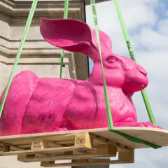 Eine große, pinke Hasenskulptur wird an grünen Gurten über einer Holzpalette aufgehängt.