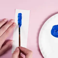 Eine Hand hält einen Pinsel, um mit leuchtendem Blau auf weißem Papier zu malen. Daneben auf einer Pappplatte wird dunkelblau gemischt.