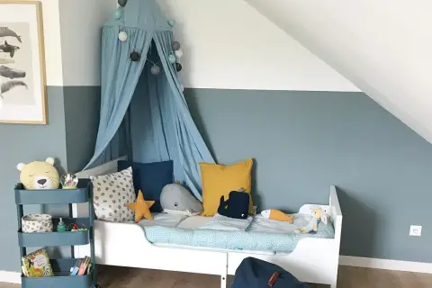 Ein Kinderzimmer, in dem blau und grau mischen ergibt eine beruhigende Wandfarbe. Ein Himmelbett mit Wal-Kissen und ein Teddybär auf einem Rollwagen.