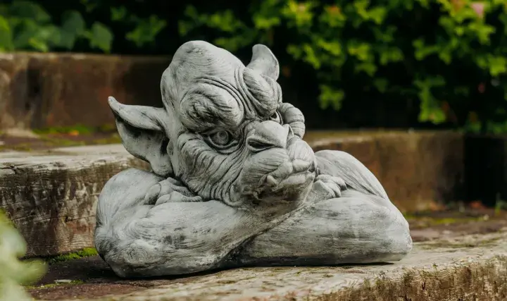 Graue Gartenskulptur eines Fabelwesens, das auf einem Holzstamm ruht, mit grünem Laub im Hintergrund.