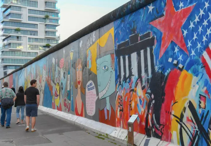 Bunte East Side Gallery in Berlin mit Graffiti und Menschen, im Hintergrund ein modernes Hochhaus.