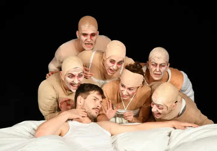 Schauspieler mit gruseliger Schminke und Glatzenköpfen umringen einen Mann im Bett. Die Szene könnte die genretypische Bedeutung von Horror oder Thriller verdeutlichen.