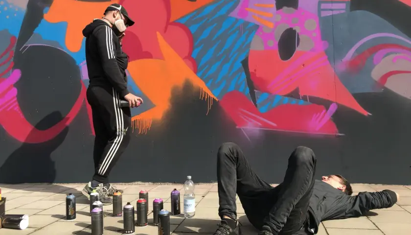 Zwei Künstler, eine Grafikdesignerin und ihr Kollege, sprühen Graffiti. Einer liegt entspannt, der andere sprüht Farbe auf die Wand.
