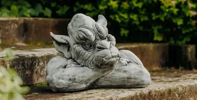 Graue Gartenskulptur eines Fabelwesens, das auf einem Holzstamm ruht, mit grünem Laub im Hintergrund.