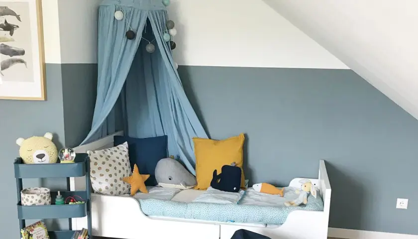 Ein Kinderzimmer, in dem blau und grau mischen ergibt eine beruhigende Wandfarbe. Ein Himmelbett mit Wal-Kissen und ein Teddybär auf einem Rollwagen.