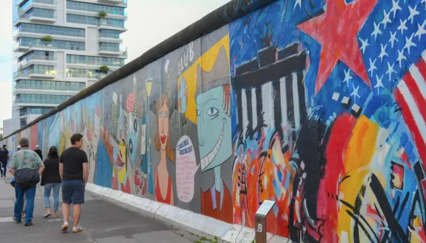 Bunte East Side Gallery in Berlin mit Graffiti und Menschen, im Hintergrund ein modernes Hochhaus.