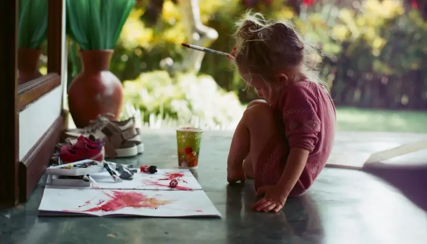 Kleines Mädchen malt mit Pinsel und Farben, um orange zu mischen. Bunte Tupfer auf Papier, ein Becher und Schuhe daneben.