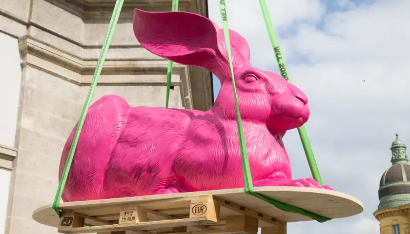 Eine große, pinke Hasenskulptur wird an grünen Gurten über einer Holzpalette aufgehängt.