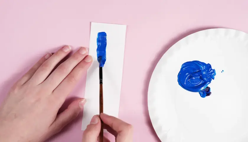 Eine Hand hält einen Pinsel, um mit leuchtendem Blau auf weißem Papier zu malen. Daneben auf einer Pappplatte wird dunkelblau gemischt.