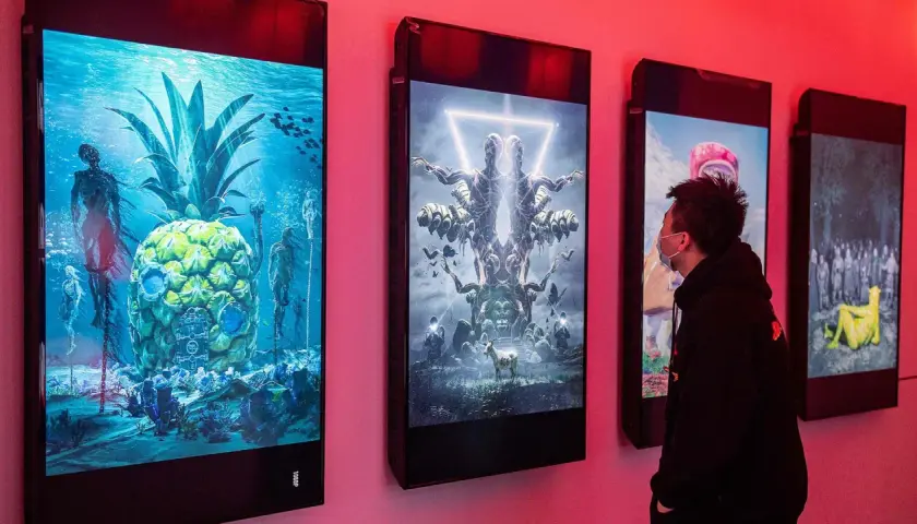 Ein Mann betrachtet digitale Kunstwerke, die an einer Wand hängen. Die Werke zeigen surreale Szenen, wie eine Ananas unter Wasser mit Skeletten und eine symmetrische Figur mit vielen Armen. Diese Werke stammen von einem berühmten Grafik Künstler.