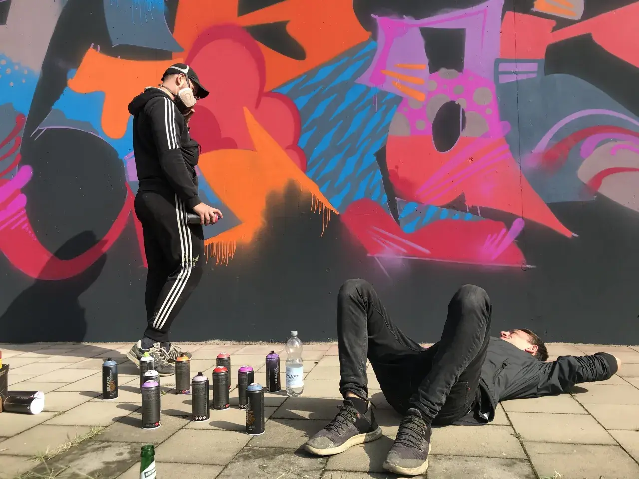 Zwei Künstler, eine Grafikdesignerin und ihr Kollege, sprühen Graffiti. Einer liegt entspannt, der andere sprüht Farbe auf die Wand.