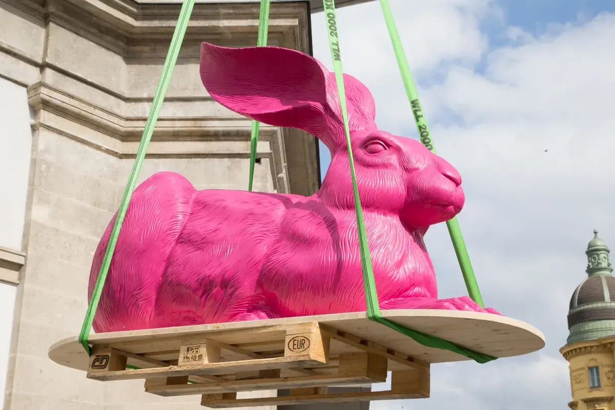 Eine große, pinke Hasenskulptur wird an grünen Gurten über einer Holzpalette aufgehängt.