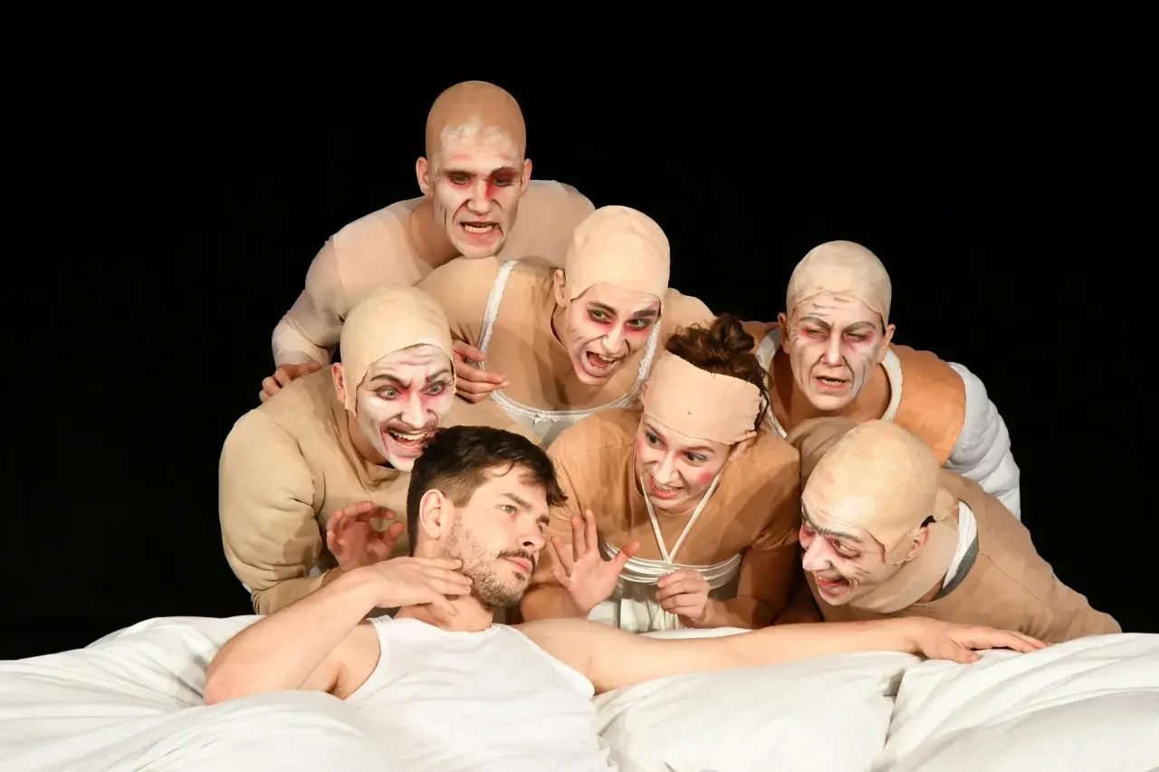 Schauspieler mit gruseliger Schminke und Glatzenköpfen umringen einen Mann im Bett. Die Szene könnte die genretypische Bedeutung von Horror oder Thriller verdeutlichen.