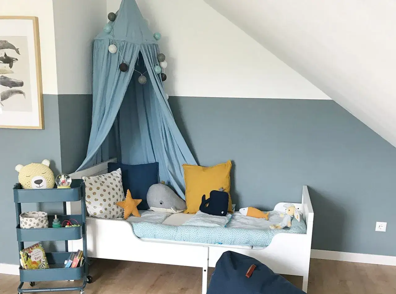 Kinderzimmer mit Himmelbett, Spielzeug und einem Rollwagen. Die Wandfarbe, wo blau und grau mischen ergibt, passt gut zu den maritimen Deko-Elementen.