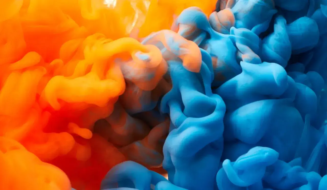 Orange und blaues Farbpulver mischen sich in Wasser zu wirbelnden Wolken.